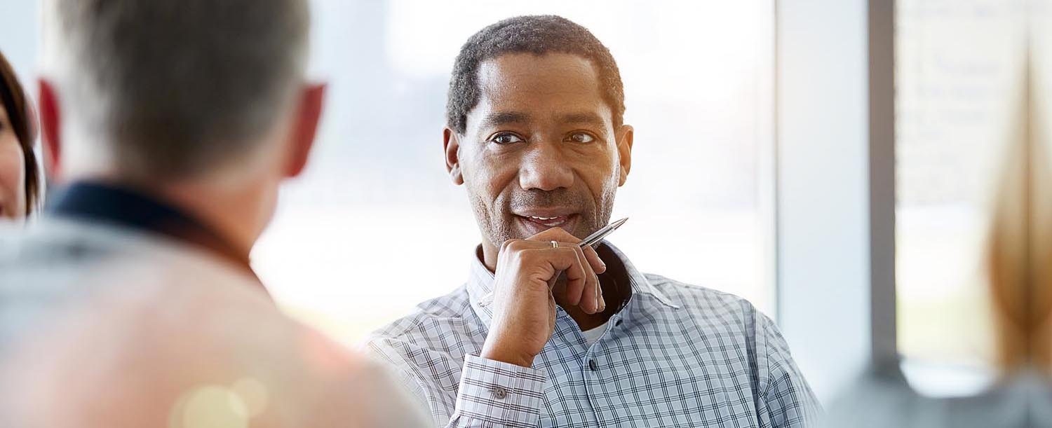 A man holds a pen while listening to another man in the foreground